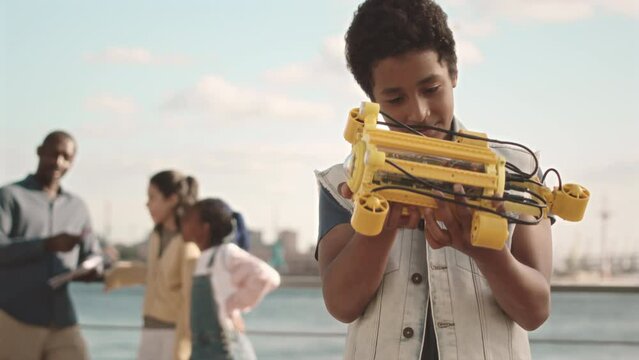 Medium slowmo portrait of African American schoolboy with hydraulic centralizer in hands smiling at camera standing at sunny quay during outdoor engineering class with teacher and classmates