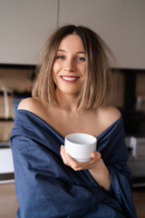 Sleepy positive woman wrapped in blue bed sheet drinking coffee in the morning in the kitchen