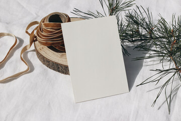 Christmas stationery. Blank greeting card, invitation mockup n cut wooden round board. Green pine tree branch and velvet ribbon in sunlight. White linen tablecloth. Festive winter still life template