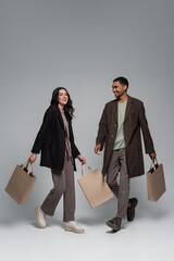 full length of happy interracial couple in stylish outfits holding shopping bags and walking on...