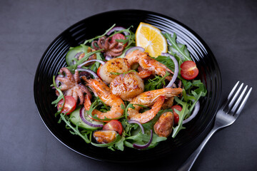 Salad with seafood, arugula, tomatoes, cucumbers, red onion and lemon on a black plate. Black background, close-up.