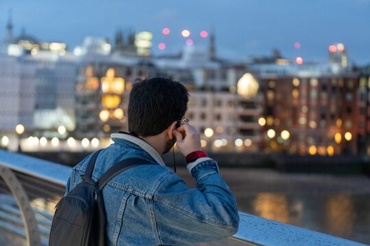Back view of a young man in earphones overlooking the ondon skyline from a bridge over the river