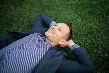 Happy Young businessman in a suit lying on the green grass and relaxing in park.