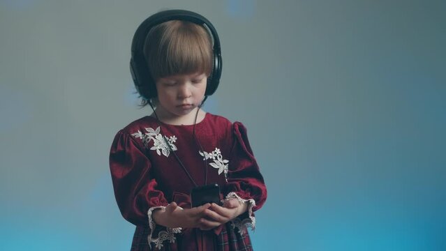 Cute little girl in big headphones and a beautiful vintage dress with lace is listening to music on her phone. There is a color music in the background. The concept of music mania and audiophilia