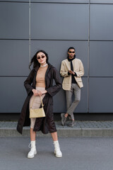 full length of stylish young woman in autumnal coat and sunglasses standing with handbag near african american man.