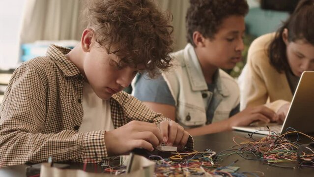 Waist up slowmo of smart Biracial schoolboy connecting wires while doing project on electronics with classmates in class