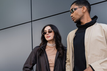 low angle view of stylish multiethnic couple in trendy autumnal outfits and sunglasses standing near building.