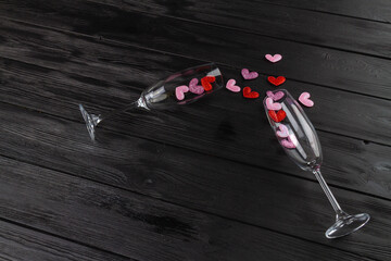Two champagne glasses with spilled sequin hearts. Black wooden desk. Happy valentines day flat lay.