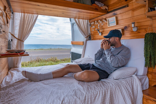 Hombre joven con gorra acostado en la cama de la camper, con la taza tomando caf&eacute;, con expresi&oacute;n de relax. Fotograf&iacute;a horizontal con copy space.