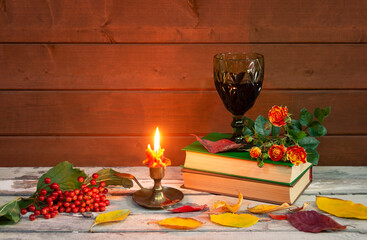Glass of red wine, burning candle, stack of books, orange rose, autumn leaves and viburnum berries on aged wooden table. Selective focus.