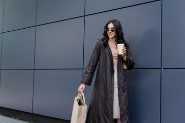 cheerful young woman in stylish outfit and sunglasses holding paper cup and shopping bag near building.