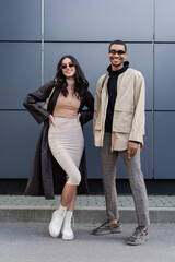full length of stylish interracial couple in autumnal outfits and sunglasses standing near building.