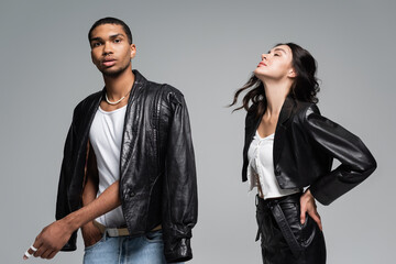pretty young woman posing with closed eyes near african american man in leather jacket isolated on grey.