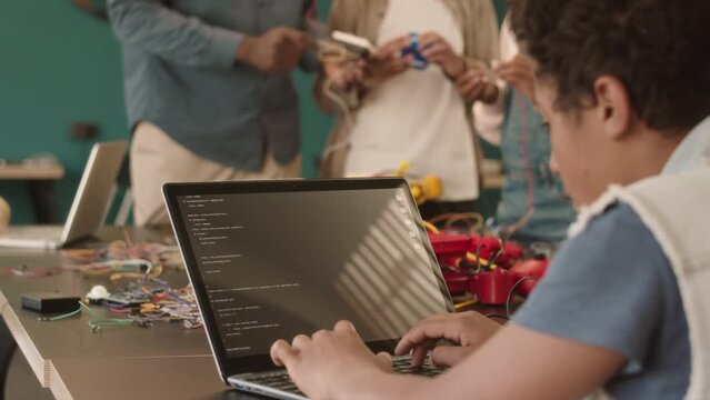 African American schoolboy writing program code on laptop while constructing robots together with teacher and classmates having class of electronics at school