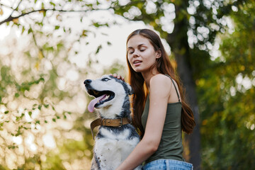 A woman with a husky breed dog smiles and affectionately strokes her beloved dog while walking in nature in the park in autumn against the backdrop of sunset