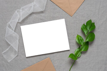 Blank invitation wedding card mockup, brown envelopes, ribbon and green leaves.Grey linen textile background. Top view, flat lay.