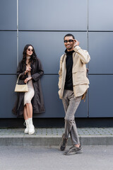 full length of happy african american man adjusting sunglasses near stylish woman standing with handbag.