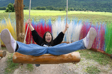 A happy Asian kid in a black shirt on the wooden swing with a background of colorful trees, grasses, and mountains in natural daylight from Thailand.