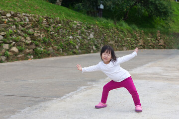 An Asian kid in a white shirt and purple pants in action with a smiley face in natural daylight. An Asian girl kid with rocks in the background.
