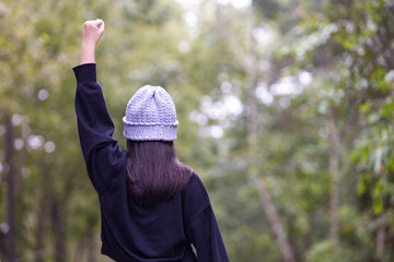 Back of a long hair Asia girl with a grey crochet hat and black shirt is raising her left hand with blur background in natural light.