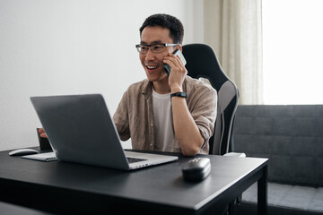 Happy adult asian man wearing glasses using laptop talking on smartphone at home. Remote job, freelance working concept.