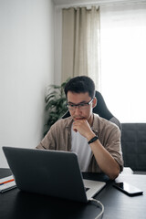 Adult asian man wearing glasses sitting at table using laptop at home. Remote job, freelance working concept.
