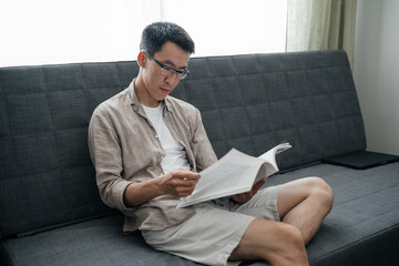 Calm adult asian man wearing glasses sitting on sofa reading book or Magazine. Having rest, education.