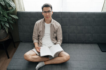 Calm adult asian man wearing glasses sitting on sofa looking at camera with book or Magazine. Having rest, education.