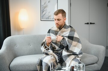Sick bearded man who has bad cold or seasonal flu sitting on couch at home. Guy with fever wearing warm plaid shivering with worried face expression.