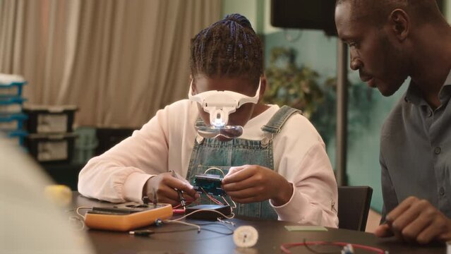Waist up slowmo of African American school girl in magnifying goggles examining small electronical part having technology and robotics class with modern African American male teacher