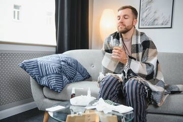 Sick bearded man who has bad cold or seasonal flu sitting on couch at home. Guy with fever wearing...