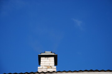 Chimney against the blue sky. The white brick chimney of the spherhu stove is covered with an iron umbrella deflector to protect it from moisture. Barely noticeable gray smoke comes out of the chimney