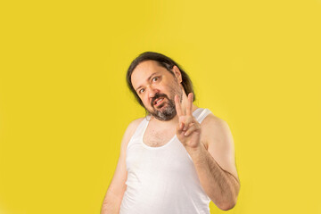 A disgruntled unshaven man in a white undershirt shows a two-finger peace gesture. Unemployed slovenly dressed man from Eastern Europe. - obrazy, fototapety, plakaty