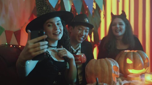 A group of friends are sitting at a table in creepy costumes, taking pictures on their phone and laughing on Halloween