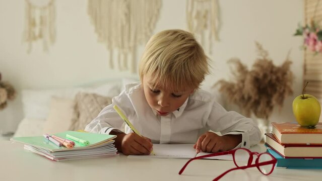 Cute preschool blond child, boy, writing on the table in notebook, apple, wearing glasses, preparing for school