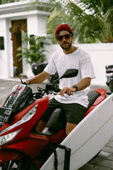 Surfer rides on motorbike with surfboard. Bali island, Indonesia. Young stylish man driving a scooter. Surfer with a surfboard on a rack on a motorcycle.