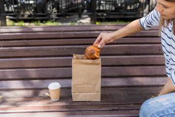 The girl is sitting on a bench in the park. Drinking coffee with a croissant. The girl takes out a croissant from the bag. Close-up of a hand holding croissant under paper bag. Sunny day in park.