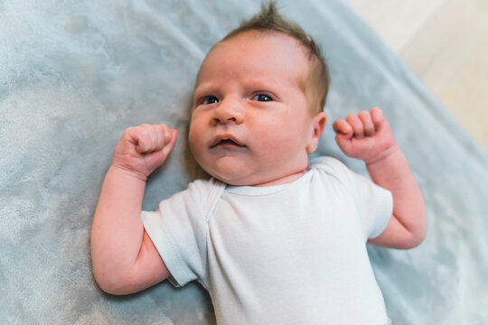 Closeup indoor portrait of a european infant baby boy in light blue bodysuit clenching his fists and giving a surprised look at camera. Funny facial expressions. High quality photo