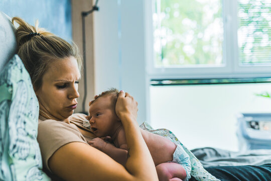 Blond white mother lying in bed looking with concerned expression at her newborn baby resting on her chest. Motherhood. Caring. Horizontal indoor shot. High quality photo