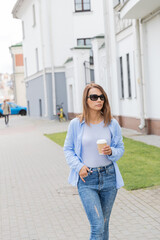 A girl walks along the street with a сup of coffee. Craft paper cup with white plastic lid. It's warm outside. The girl is dressed in jeans and blue blouse. Street view on background.