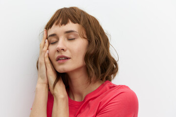 Fototapeta premium a gentle, relaxed, sleepy woman stands in a red T-shirt on a white background and holds her hands folded in her palms under her head, closing her eyes