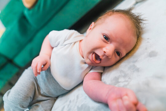 Funny facial expressions by newborns. Caucasian infant baby boy opening his mouth and sticking his tongue out while looking at camera and lying down on gray mattress. High quality photo