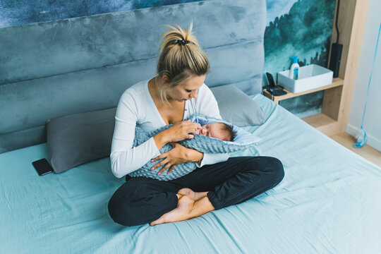 Caucasian blonde mother sitting in bed with her baby boy wrapped in blanet in her lap touching his face affectionately and looking at him. Caring, Horizontal indoor shot. High quality photo