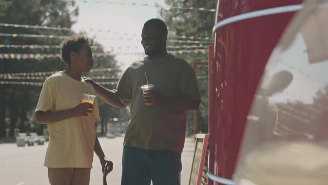 Medium shot of African American man and his 12 year old son buying refreshing drinks at street food truck, chatting and having good time, spending weekends together in park on sunny day