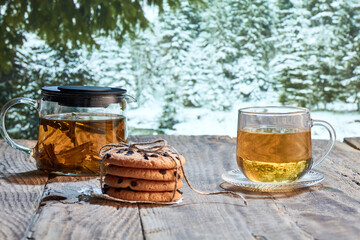 Winter still life. Glass pot with herbal tea, cakes, biscuits on wooden table over winter snow forest background. Design for greeting card, wallpaper for device screen