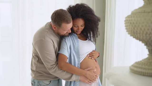 White man touching belly of his pregnant black wife. Pregnant woman and loving husband hugging tummy at home. Feeling baby movement by multiethnic couple on pregnant belly. 