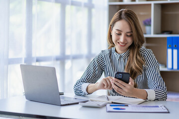 Asian businesswoman in a formal suit in the office is happy and cheerful while using a smartphone to work, financial accounting, analytical concept.
