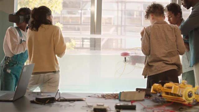 Rear view slowmo of group of diverse schoolchildren and their modern teacher of robotics experimenting with red robo boat floating in aquarium in school lab