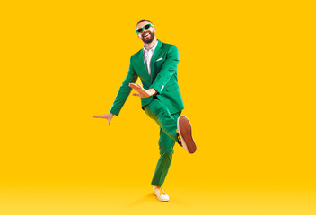 Funny young guy in a party suit dancing in a modern studio. Full body shot of a happy, cheerful, goofy man wearing a green suit and sunglasses dancing isolated on a bright yellow colour background - obrazy, fototapety, plakaty