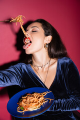 sexy woman in blue velour dress eating tasty spaghetti on red background.